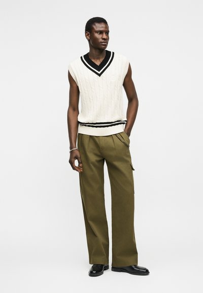 Άνδρας που στέκεται με το αριστερό χέρι στην τσέπη, φοράει λευκό πλεκτό αμάνικο πουλόβερ με μαύρο περίγραμμα, παντελόνι ελιάς φαρδύ στο πόδι και μαύρα παπούτσια.