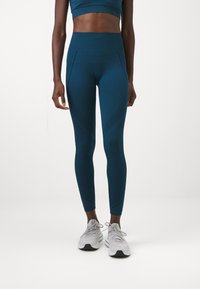 Teal taille-hoge legging met textuurdetails en naadaccenten, gecombineerd met grijze sportschoenen. Rekbare, aansluitende pasvorm.