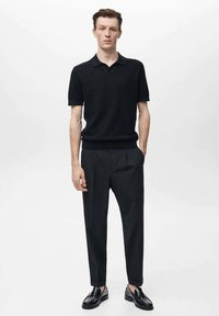 Polo shirt nero a maglia con maniche corte, abbinato a pantaloni neri su misura. Il modello è in piedi contro uno sfondo bianco semplice, indossando scarpe nere.