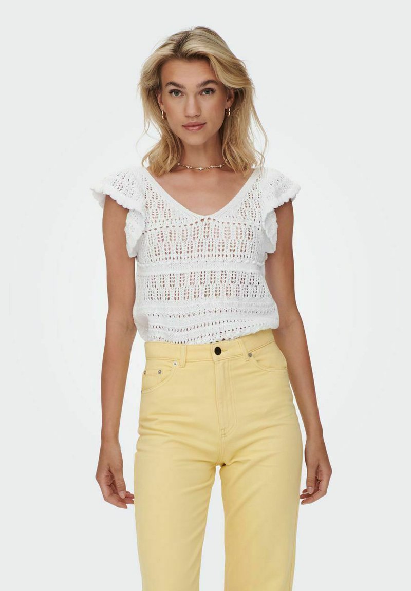 Top de punto blanco con mangas cortas y volantes, que presenta un patrón de punto abierto. Combinado con jeans amarillos de cintura alta.