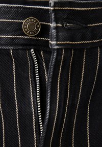 Gros plan sur un tissu noir à fines rayures avec un bouton en métal et une fermeture éclair partiellement ouverte sur un pantalon ou un jean.
