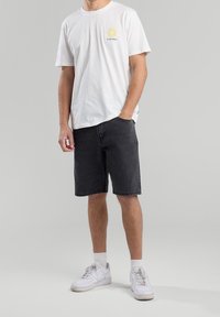 Mies, joka on pukeutunut valkoiseen T-paitaan, jossa on keltainen kukka ja teksti "California", mustiin farkkushortseihin, valkoisiin sukkiin ja valkoisiin tennareihin, seisoo käsi taskussa.