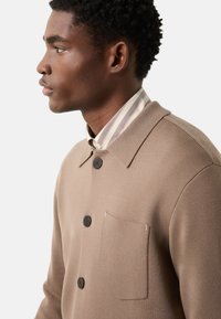 Braune gestrickte Jacke mit schwarzen Knöpfen, einer Brusttasche und einem Kragen über einem gestreiften Hemd. Glatte Textur und lässiges Design.