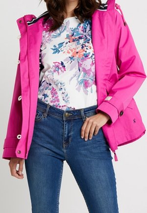 Kvinde iført en knaldrød regnjakke over en hvid blomstret top og blå jeans, med den ene hånd hvilende på hoften.