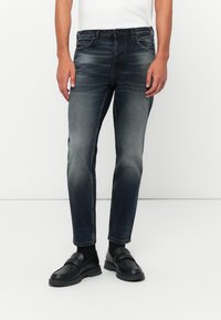 Vaqueros de mezclilla de un azul marino con corte slim, lavado desgastado, cinco bolsillos y costuras en contraste. Combinados con mocasines negros y una camisa blanca.