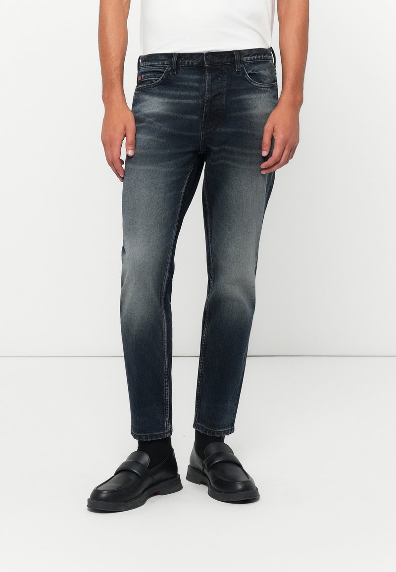 Vaqueros de mezclilla de un azul marino con corte slim, lavado desgastado, cinco bolsillos y costuras en contraste. Combinados con mocasines negros y una camisa blanca.