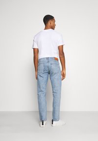 Calças de denim azul claro com corte reto, apresentando dois bolsos traseiros e um patch de marca em pele castanha na cintura. Usadas com uma t-shirt branca.