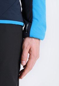 Blå och svart Ventrix-jacka visar textilstruktur på ärmarna, med distinkta färgblock och varumärkesbokstäver längs ärmslutet.