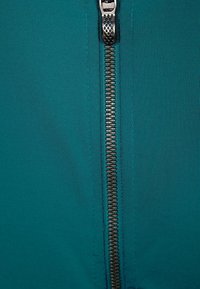 Tyrkysová látka s hladkým povrchem má vertikálně umístěný kovový zip s viditelnými zuby a dekorativním táhlem na zip nahoře.