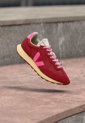 Rode en roze sneaker met beige zool zwevend boven een betonnen oppervlak met trappen op de achtergrond, gestrikt met roze veters.