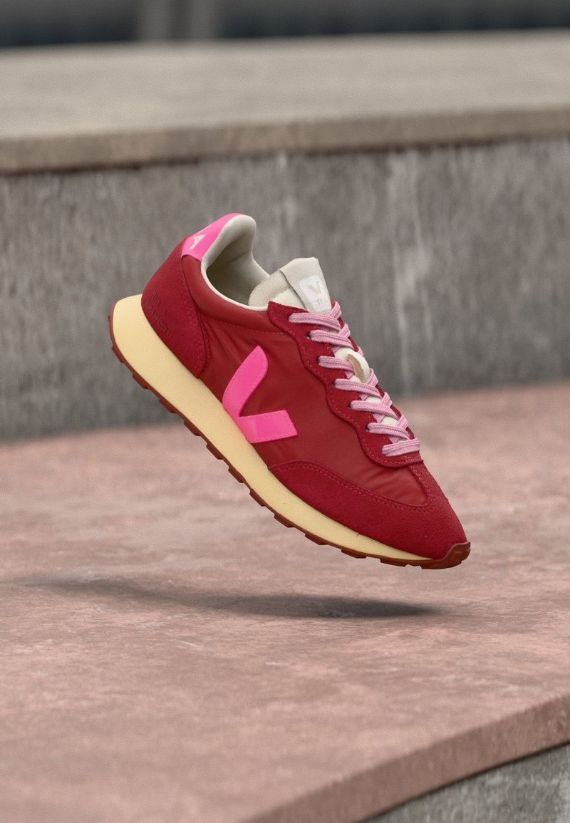 Κόκκινο και ροζ αθλητικό παπούτσι με μπεζ σόλα, αιωρούμενο πάνω από μια επιφάνεια από σκυρόδεμα και σκαλοπάτια στο φόντο, με ροζ κορδόνια.