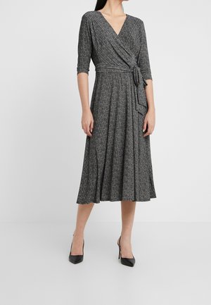 Femme portant une robe portefeuille à motifs noir et blanc avec des manches trois-quarts et des chaussures à talons noirs sur un fond uni.