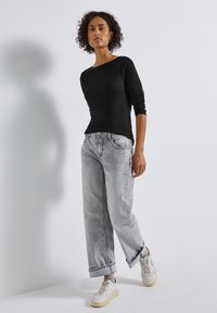 Schwarzes Langarmoberteil, kombiniert mit hellgrauen, lässigen Jeans mit umgeschlagenem Saum und Zwei-Knopf-Verschluss, getragen mit weißen und grauen Sneakers.