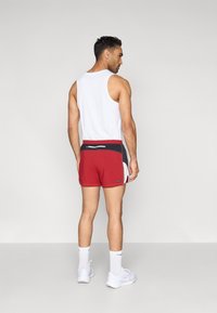 Κόκκινα αθλητικά σορτς με σκούρες μπλε και λευκές λεπτομέρειες, ελαστική μέση και στενή γραμμή, συνδυασμένα με λευκό αμάνικο τοπ. Λευκά αθλητικά παπούτσια.