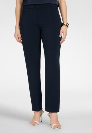 Vrouw draagt slanke donkerblauwe broek en beige sandalen met hoge hakken, staat met ontspannen handen langs het lichaam tegen een witte achtergrond.