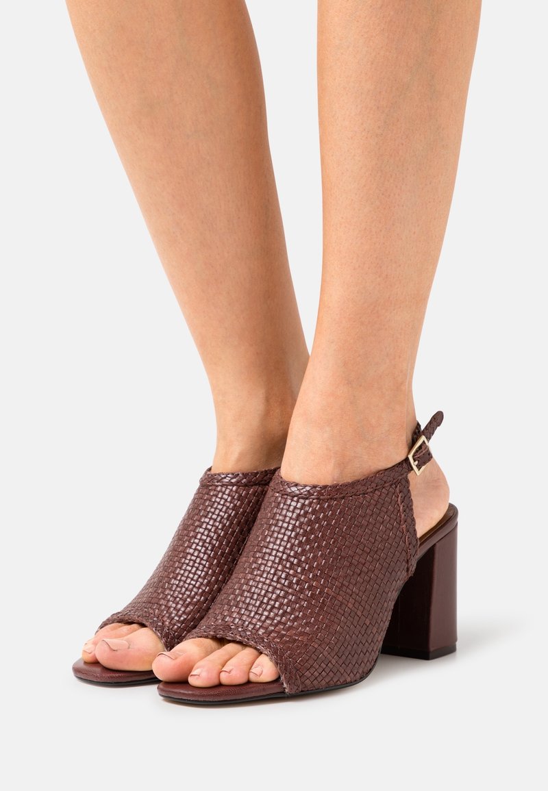 Sandales à talons ouvertes en cuir tressé marron avec brides à la cheville fixées par des boucles dorées sur pieds nus sur un fond uni.