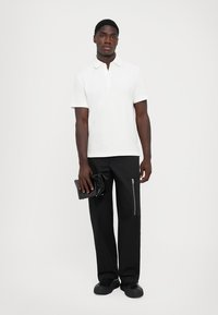 Hvit bomullspolo, svarte bukser med glidelåslommer, og en svart clutch. Modellen står mot en enkel bakgrunn.
