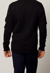 Svart stickad tröja med långa ärmar, ribbade muddarna och rundad halsringning. Verkar tight passande med en strukturerad yta. Bärs över marinblå byxor.