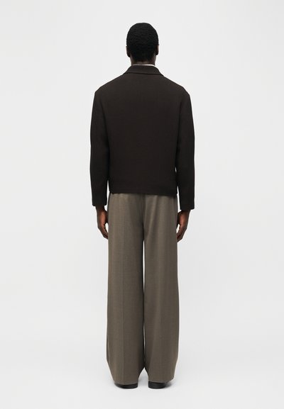 Άνδρας στέκεται πλάτη, φοράει μαύρο υφασμάτινο μπουφάν και φαρδιά γκρι παντελόνια μπροστά σε απλό φωτεινό φόντο.