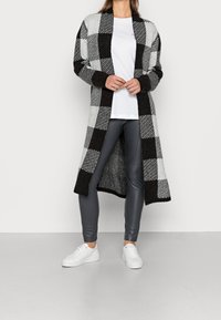 Lange gebreide cardigan in een zwart-grijs ruitpatroon, gecombineerd met grijze leggings en witte sneakers. Zachte textuur, open voorkant.