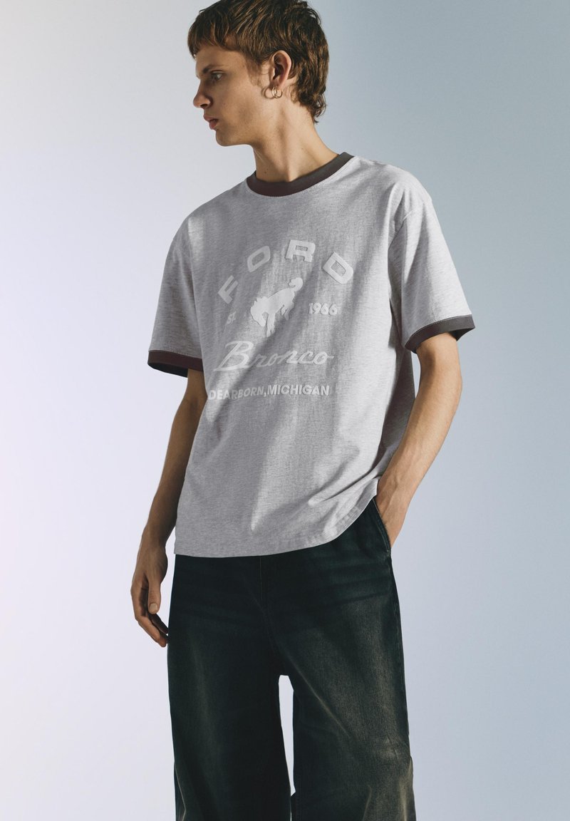 Jeune homme aux cheveux bruns portant un t-shirt gris Ford Bronco et un pantalon noir, regardant vers la gauche avec une main dans la poche.