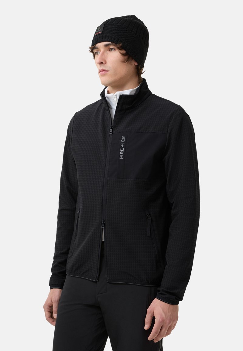 Schwarze Zip-Jacke mit einem strukturierten Muster, ausgestattet mit einer schwarzen Brusttasche, Seitentaschen und einem Stehkragen. Kombiniert mit einer schwarzen Strickmütze.