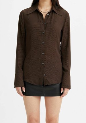 Donna che indossa una camicia marrone a fantasia con bottoni e maniche lunghe e una minigonna nera di pelle su sfondo bianco.