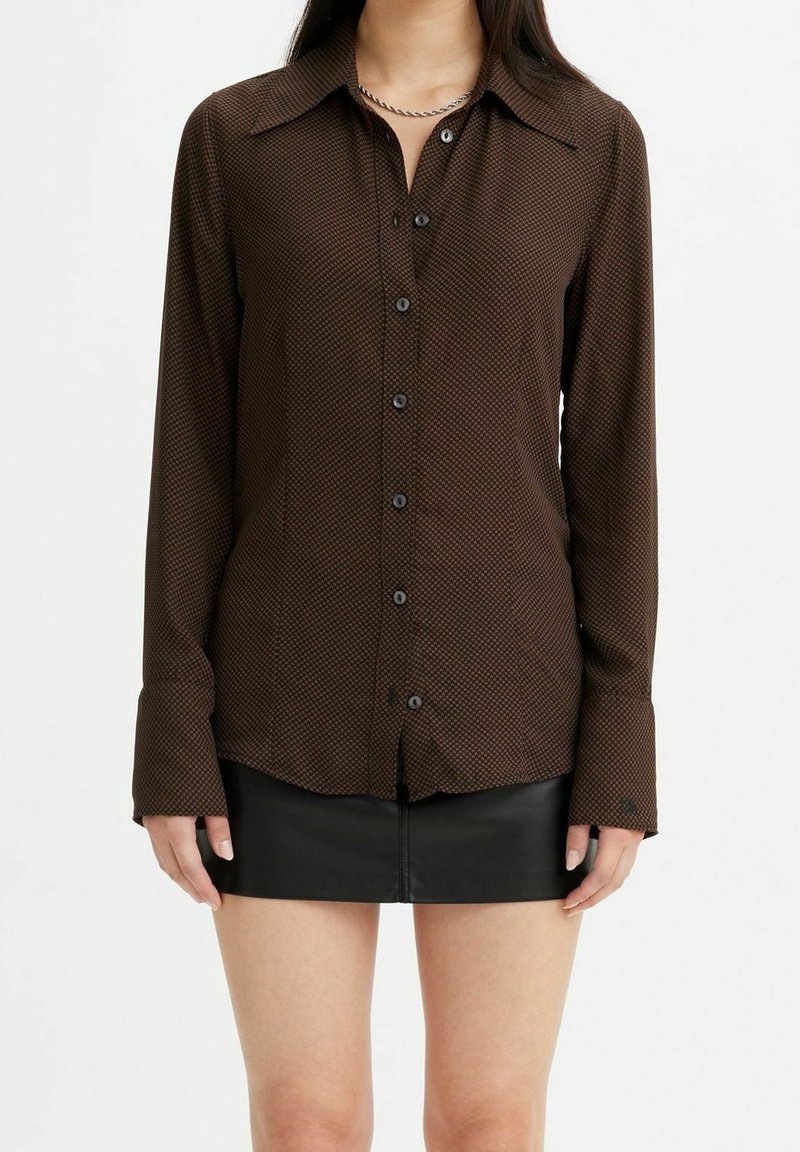 Femme portant une chemise marron à motifs avec boutons et manches longues, ainsi qu'une mini-jupe noire en cuir sur fond blanc.