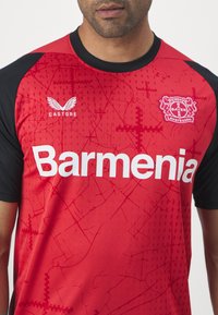 Camisa de futebol vermelha com mangas pretas. Apresenta o texto branco "Barmenia", o logótipo "CASTORE" e o emblema do "Bayer Leverkusen" com um design padrão.