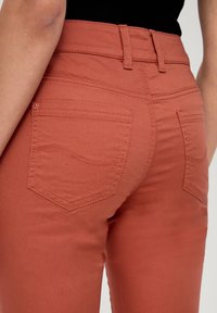 Jeans attillati color ruggine con una texture liscia, dotati di due tasche posteriori e dettagli di cucitura lungo i fianchi.