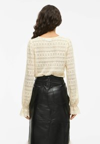 Vrouw met lang krullend haar, gekleed in een crèmekleurige kanten top met lange mouwen en een hoge zwarte leren rok, gezien van achteren.