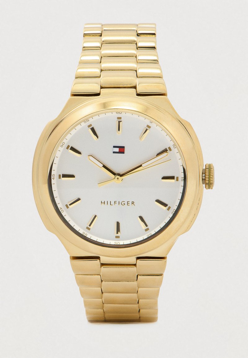 Kuldmetallist käekell valge taustaga, kuldsed tunni- ja minutimarkerid ning -näidikud, Tommy Hilfigeri logo ja seotud käevõru rihm.