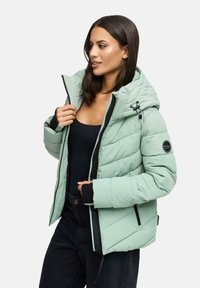 Woman wearing a light green puffer jacket with hood, adjusting the zipper, paired with black top and pants, against a plain background.