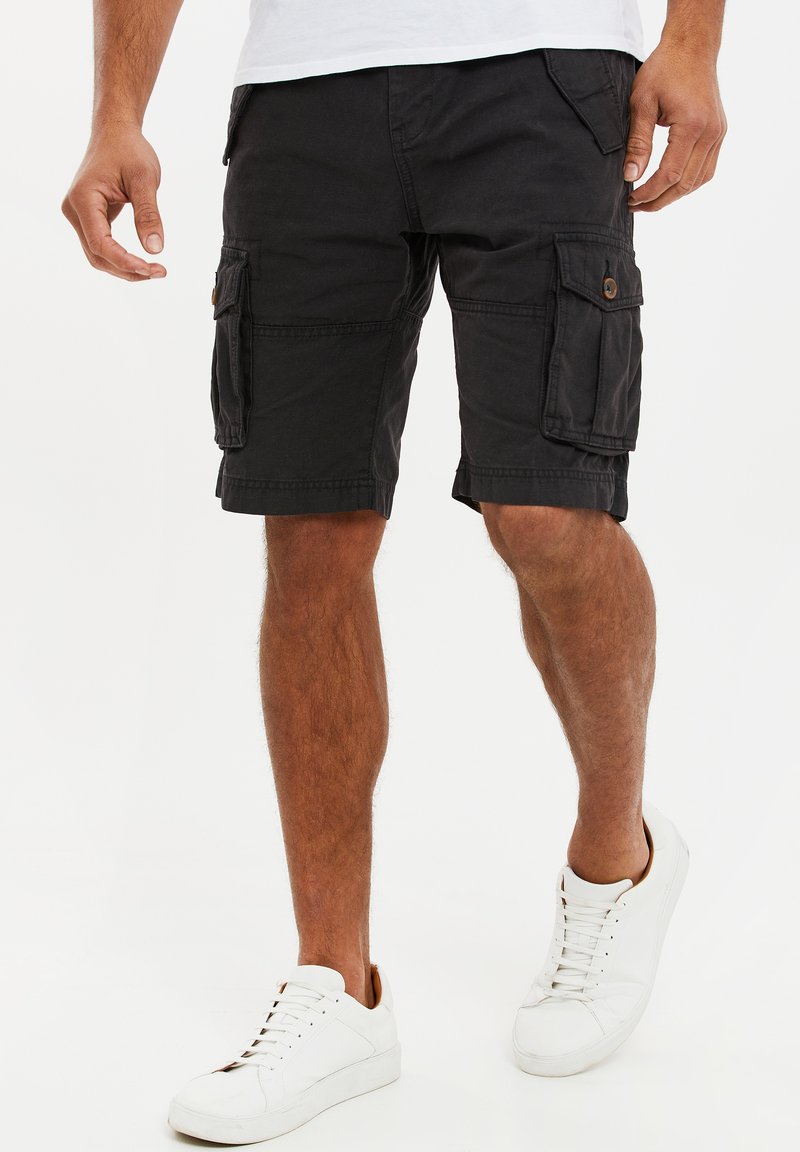 Pantalones cortos cargo negros con múltiples bolsillos, hechos de una tela duradera. Incluyen un corte relajado y una longitud hasta la rodilla, combinados con zapatillas blancas.