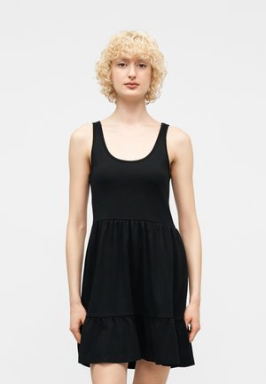 Osoba o krótkich, kręconych blond włosach, ubrana w czarną sukienkę bez rękawów z warstwami, na jednolitym jasnym tle.