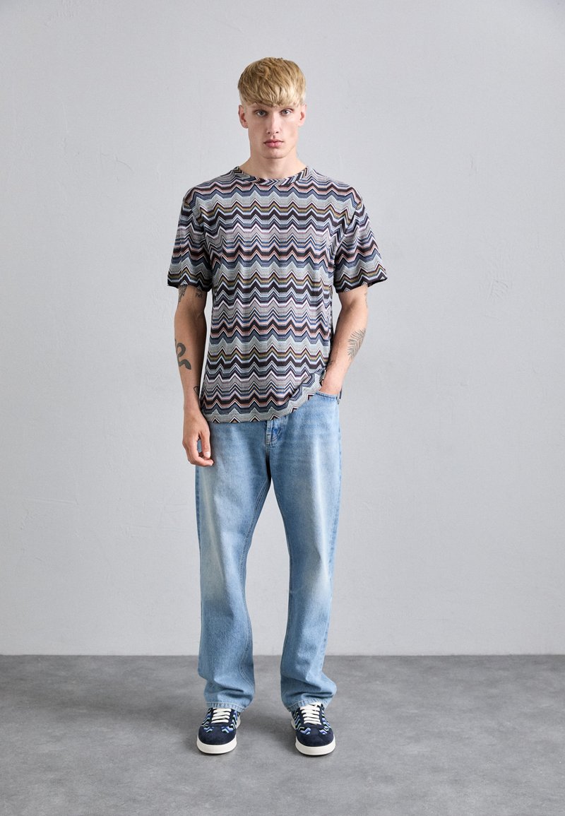 Giovane uomo con capelli biondi che indossa una maglietta a zigzag multicolore, jeans azzurri e sneakers blu navy, in piedi contro un muro grigio.