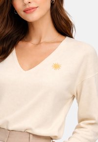 Femme portant un pull en V de couleur crème avec un petit soleil jaune brodé sur la poitrine, assorti à un pantalon beige et des boucles d'oreilles créoles dorées.