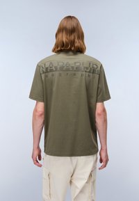 Olivengrønn bomulls t-skjorte med korte ermer, med en stor svart logo på baksiden. Designet er enkelt med en avslappet passform.