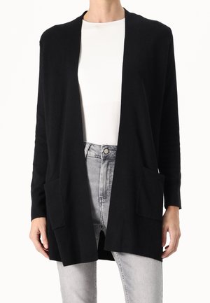 Kobieta ubrana w czarny kardigan z otwartym przodem i dwoma przednimi kieszeniami, na białej bluzce i jasnoszarych dżinsach.