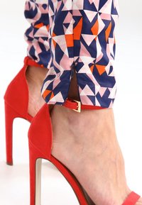 Chaussure à talon haut en daim rouge avec un nez pointu et une sangle de cheville ornée d'une boucle dorée. Un tissu à motifs géométriques colorés est visible.