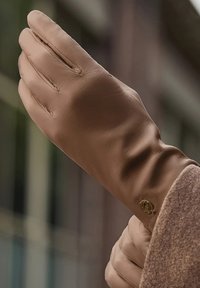 Brauner Lederhandschuh mit enger Passform, sichtbaren Nahtdetails und einem runden metallischen Logoakzent am Handgelenk. Weiche Textur offensichtlich.