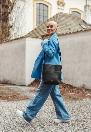 Schwarze Leder-Umhängetasche mit einfachem Design, ausgestattet mit einem silbernen Reißverschluss, die von einer Person in einer blauen Jacke und weit geschnittenen Jeans getragen wird.