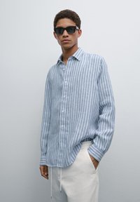 Light blue striped shirt in lightweight fabric, button-up design with a collar, long sleeves, worn with cream-colored pants. Black sunglasses.