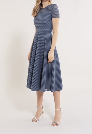 Vrouw draagt een blauw midi-jurk met korte mouwen en een diagonale strepenkant, gecombineerd met zilveren hoge hakken.