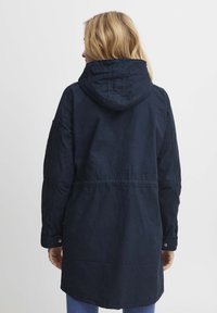 Jūras zilas kapuces jaka ar sašaurinātu jostasvietu, pogām aizdarām un nedaudz garāku aizmugures apakšdaļu. Izgatavota no teksturēta auduma ar redzamām šuvēm.