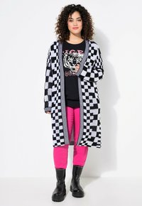 Cardigan à motif écossais noir et blanc avec une ouverture devant. Porté sur un t-shirt noir avec un graphisme de tigre et un pantalon rose. Bottines noires à la cheville.