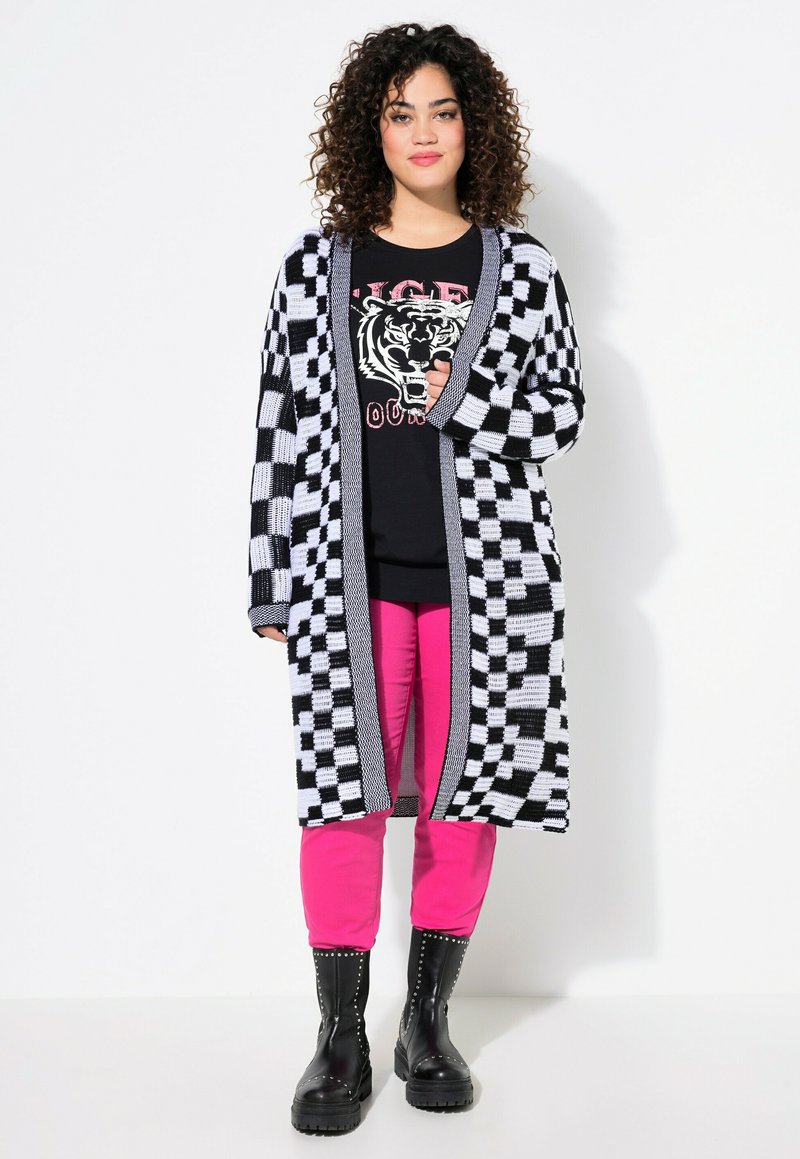 Cardigan à motif écossais noir et blanc avec une ouverture devant. Porté sur un t-shirt noir avec un graphisme de tigre et un pantalon rose. Bottines noires à la cheville.
