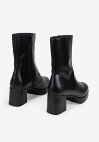 Botas de tobillo de cuero negro con una textura suave, cierre de cremallera y un tacón grueso, similar a la madera. Diseño minimalista con líneas limpias.