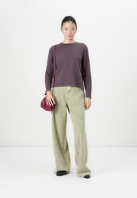 Vrouw staat gekleed in een donkerpaarse lange mouwen top, losse lichtgroene broek, zwarte schoenen en houdt een kleine bordeauxrode schoudertas vast.