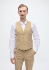 Vesta bej cu un finisaj texturat, cinci nasturi și două buzunare frontale, purtată peste o cămașă albă. Design slim fit.